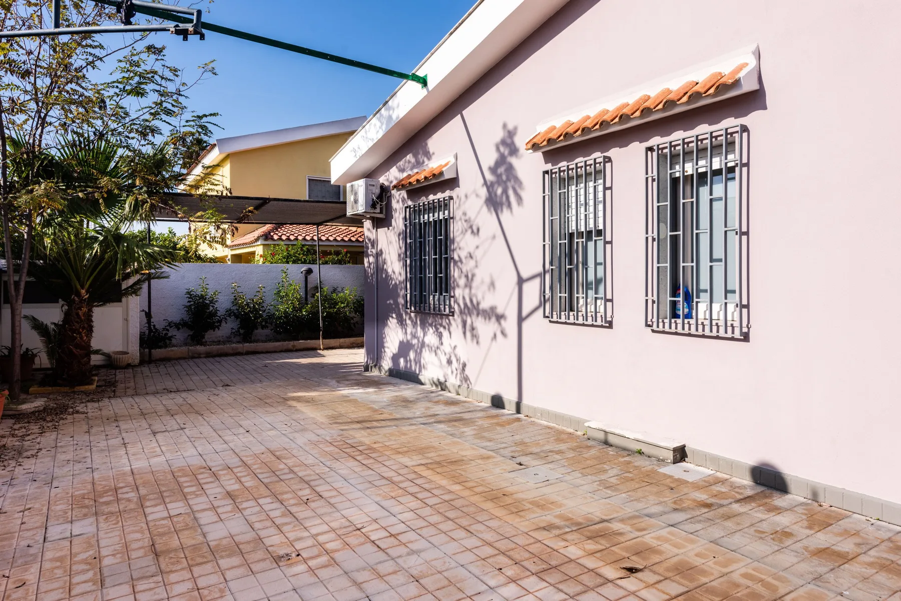 Un patio pavimentado junto a un edificio claro con ventanas enrejadas y árboles.