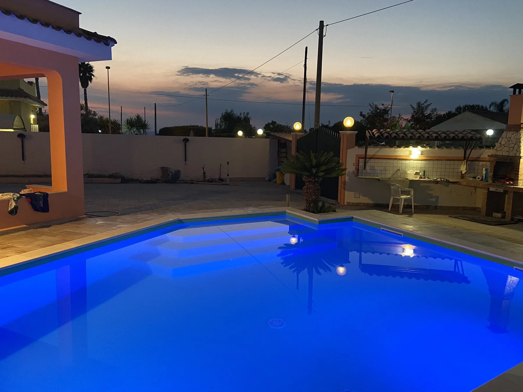 Una piscina al aire libre brilla en azul junto a un patio pavimentado al anochecer.