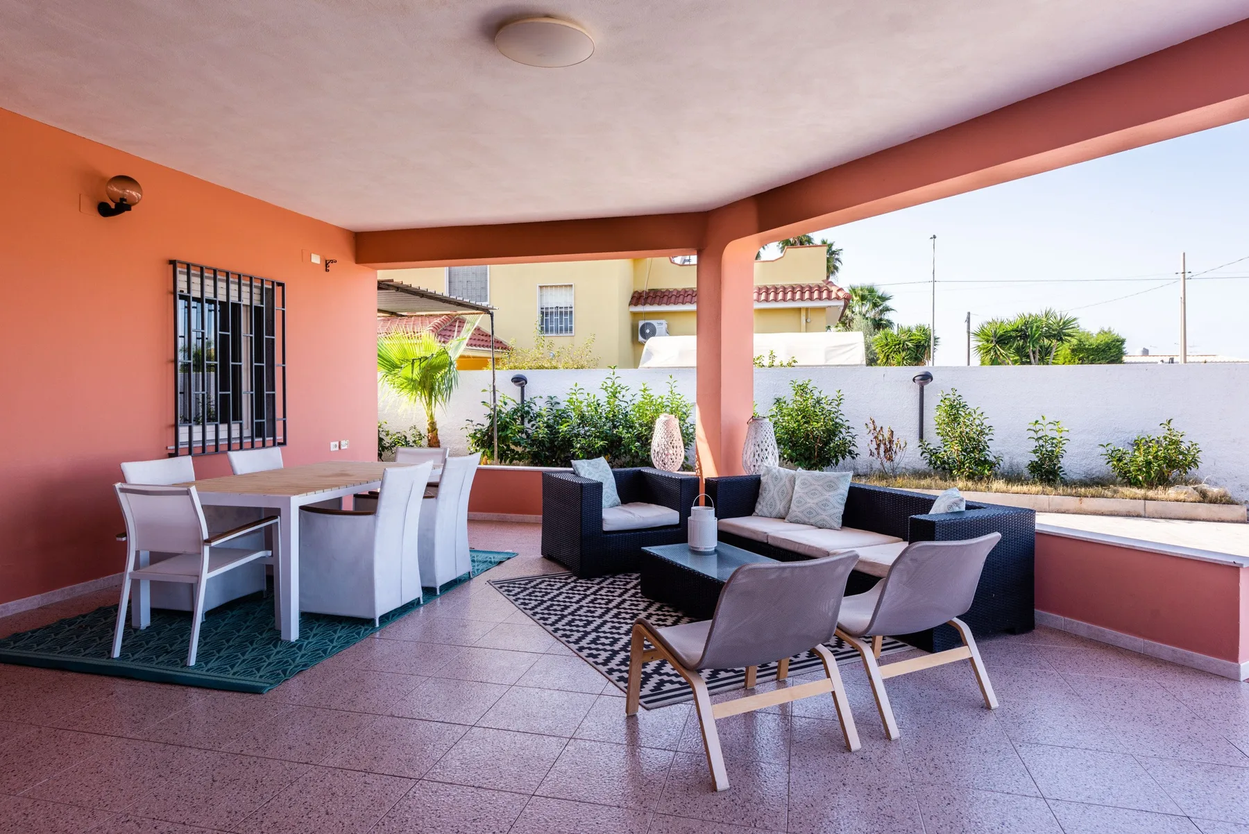 Un patio exterior cubierto con zona de comedor y asientos junto a un borde con plantas.