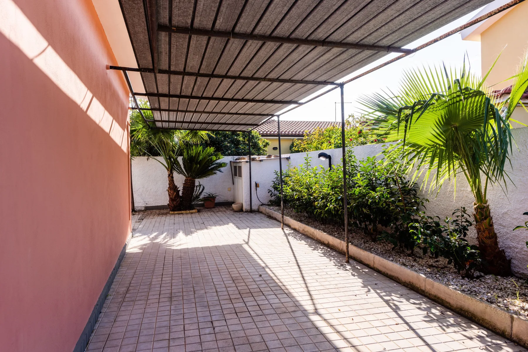 Un patio exterior pavimentado está bajo un toldo metálico, junto a plantas y una pared rosa.