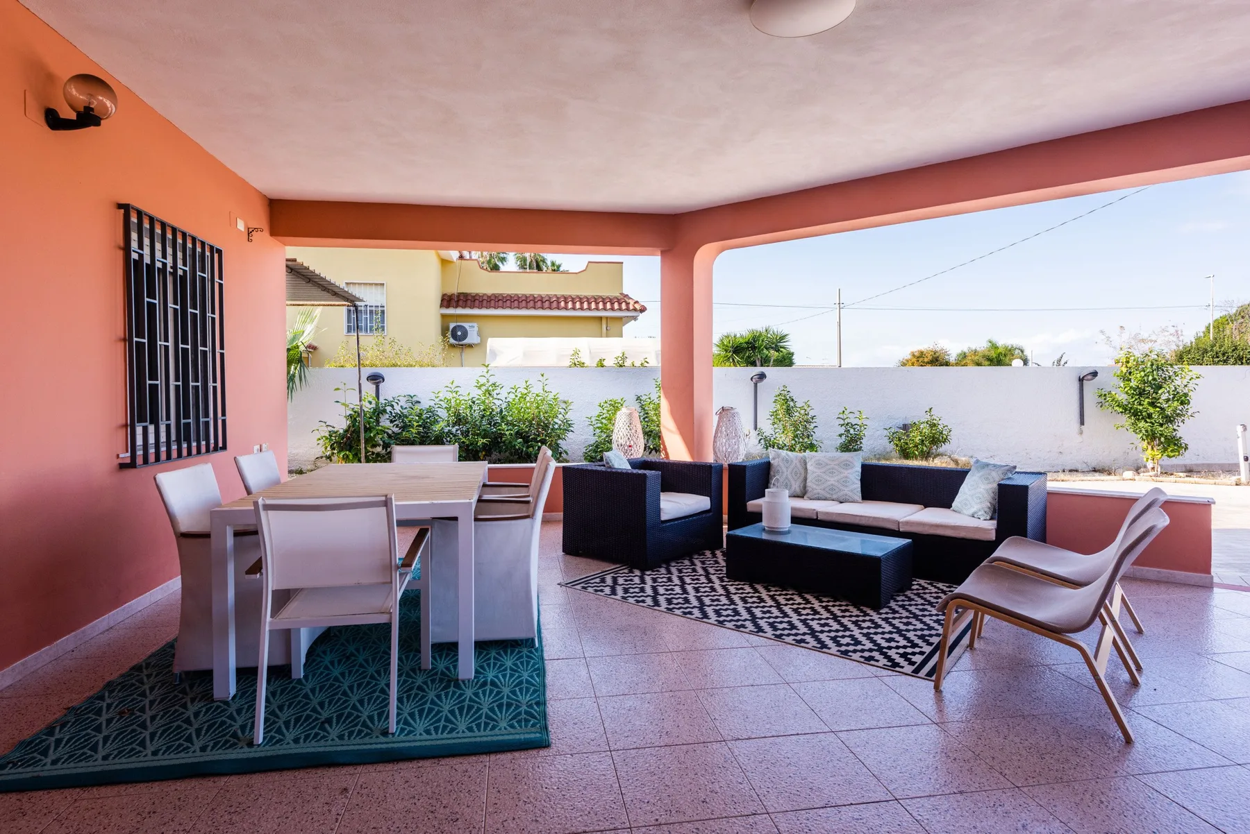 Un patio cubierto tiene un comedor y asientos de descanso junto a un muro blanco bajo.