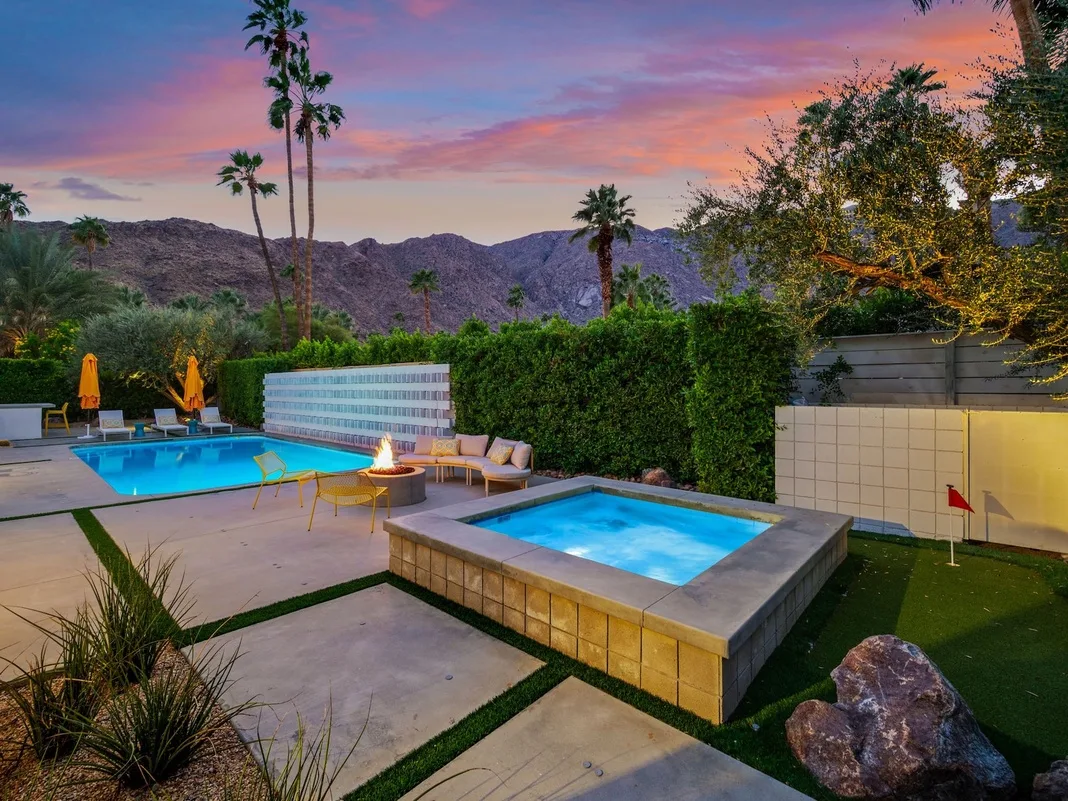 Una piscina exterior y un spa elevado están junto a asientos bajo un cielo de atardecer colorido.