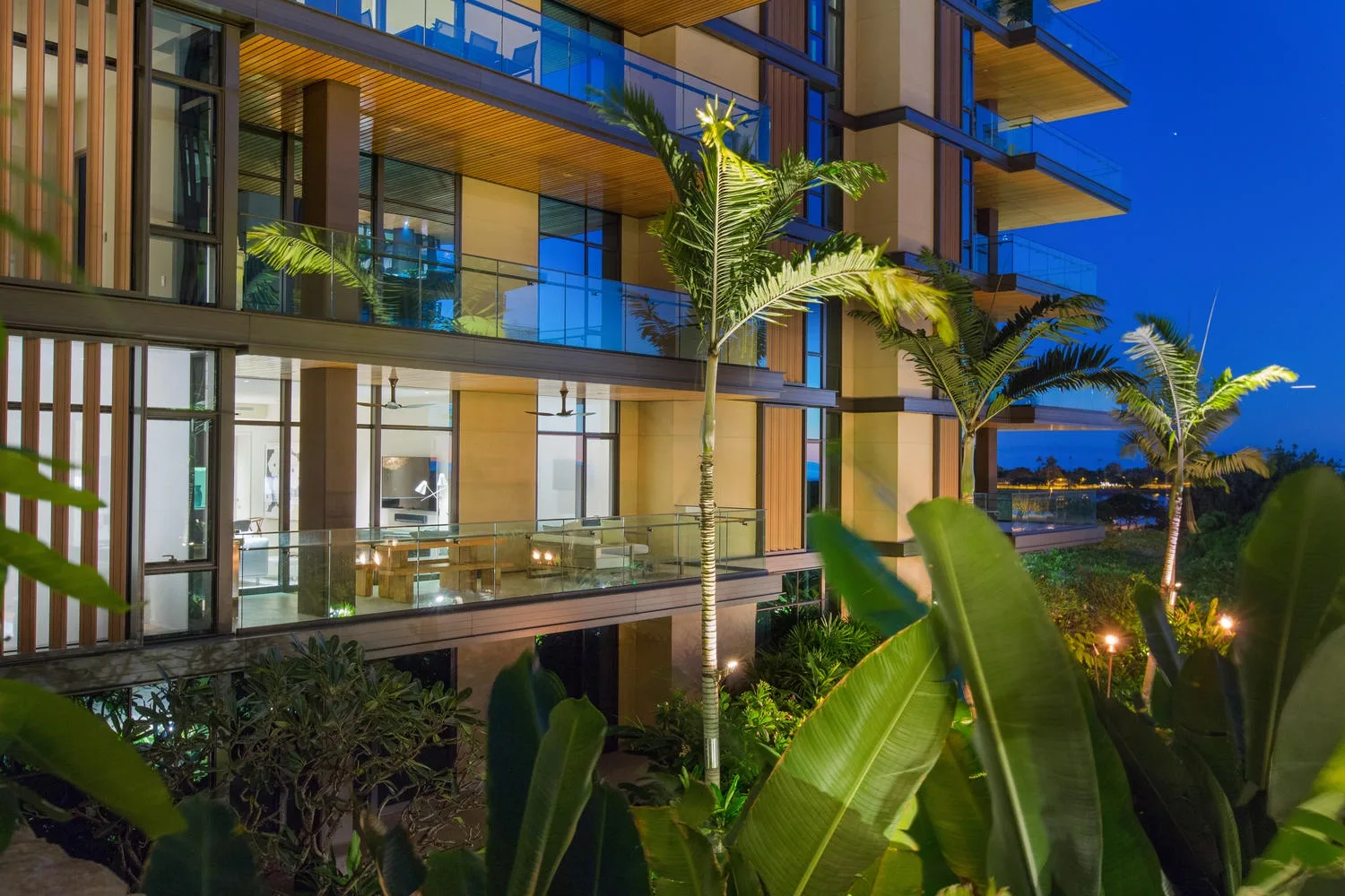 Un edificio moderno con fachada de vidrio al anochecer, rodeado de plantas tropicales.