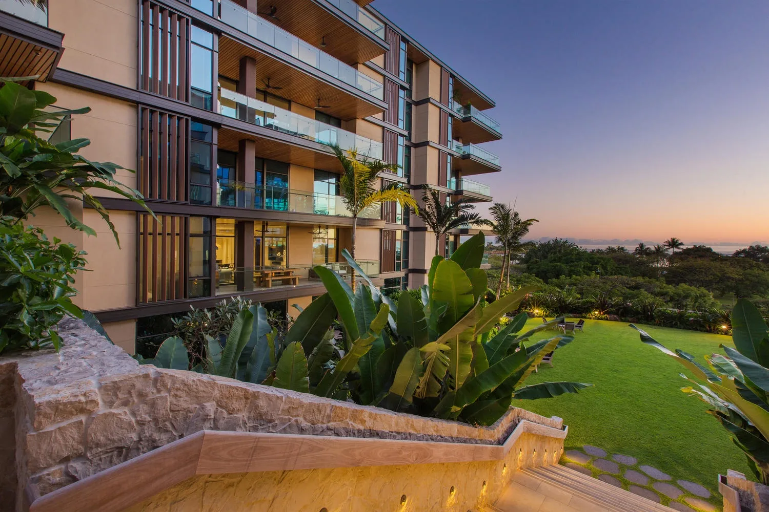 Un edificio de apartamentos moderno da a un césped ajardinado y un jardín al anochecer.