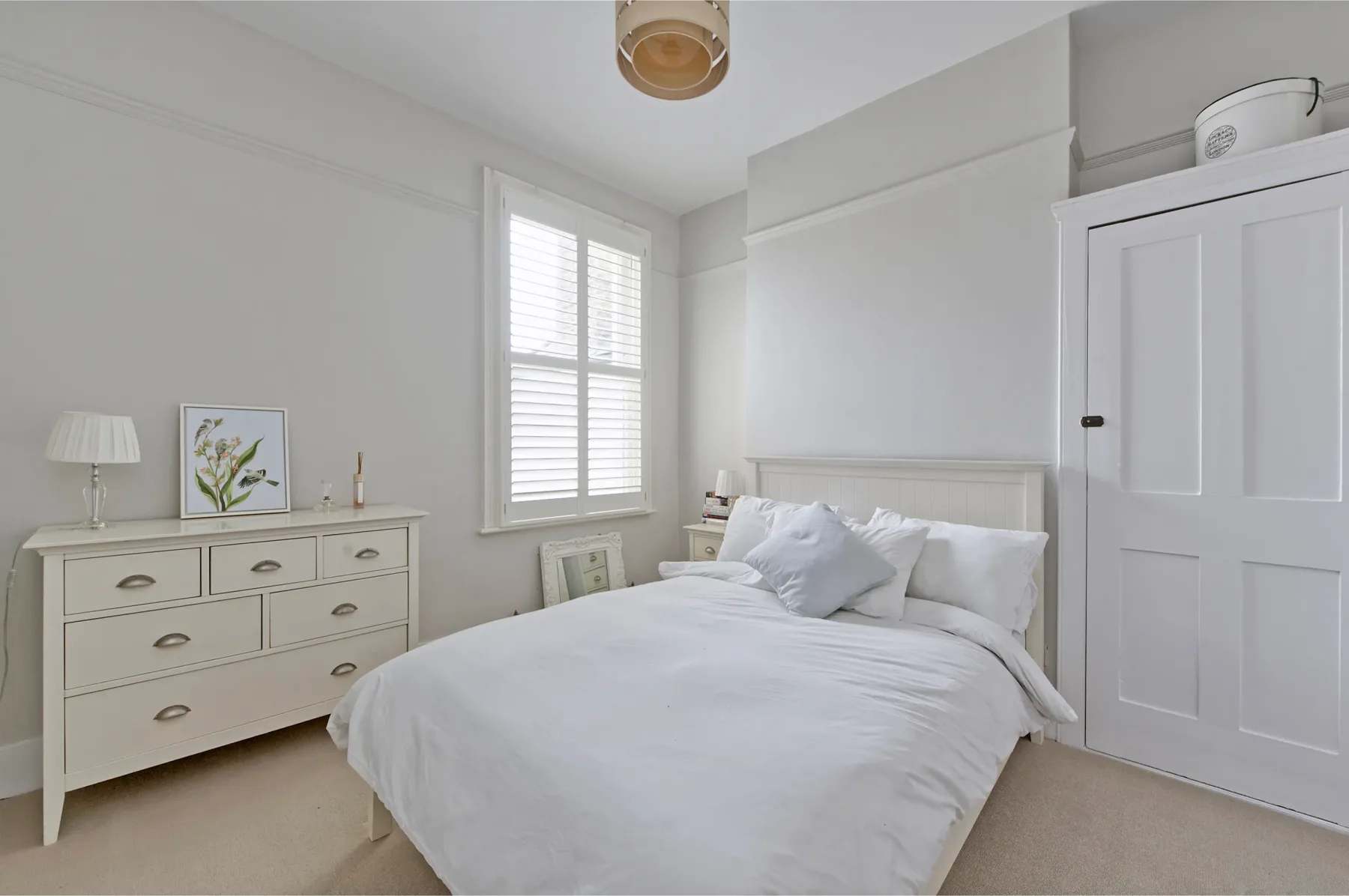 A bright bedroom with a white bed, dresser, and shuttered window.