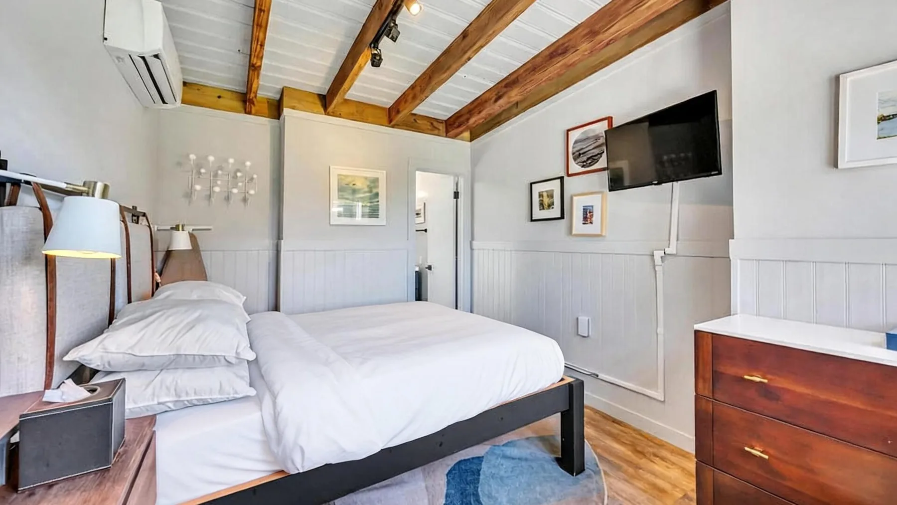 Un dormitorio luminoso con ropa de cama blanca, vigas expuestas y un televisor en la pared.