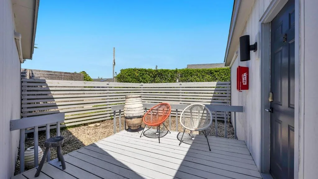 Una pequeña terraza exterior con asientos, una cerca de listones horizontales y cielo azul despejado.