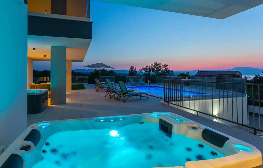 An outdoor hot tub overlooks a pool and seating area at dusk.