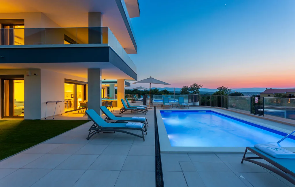 A modern house opens onto a swimming pool and patio at sunset.