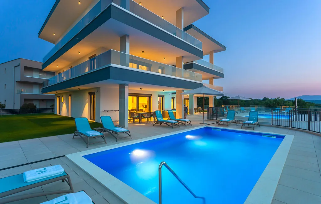A modern building overlooks a lit swimming pool and outdoor seating at dusk.