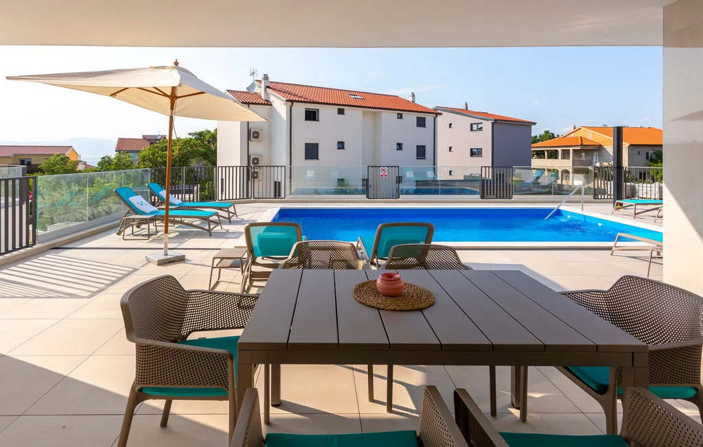 A poolside patio has an outdoor dining table, seating, and a large umbrella.