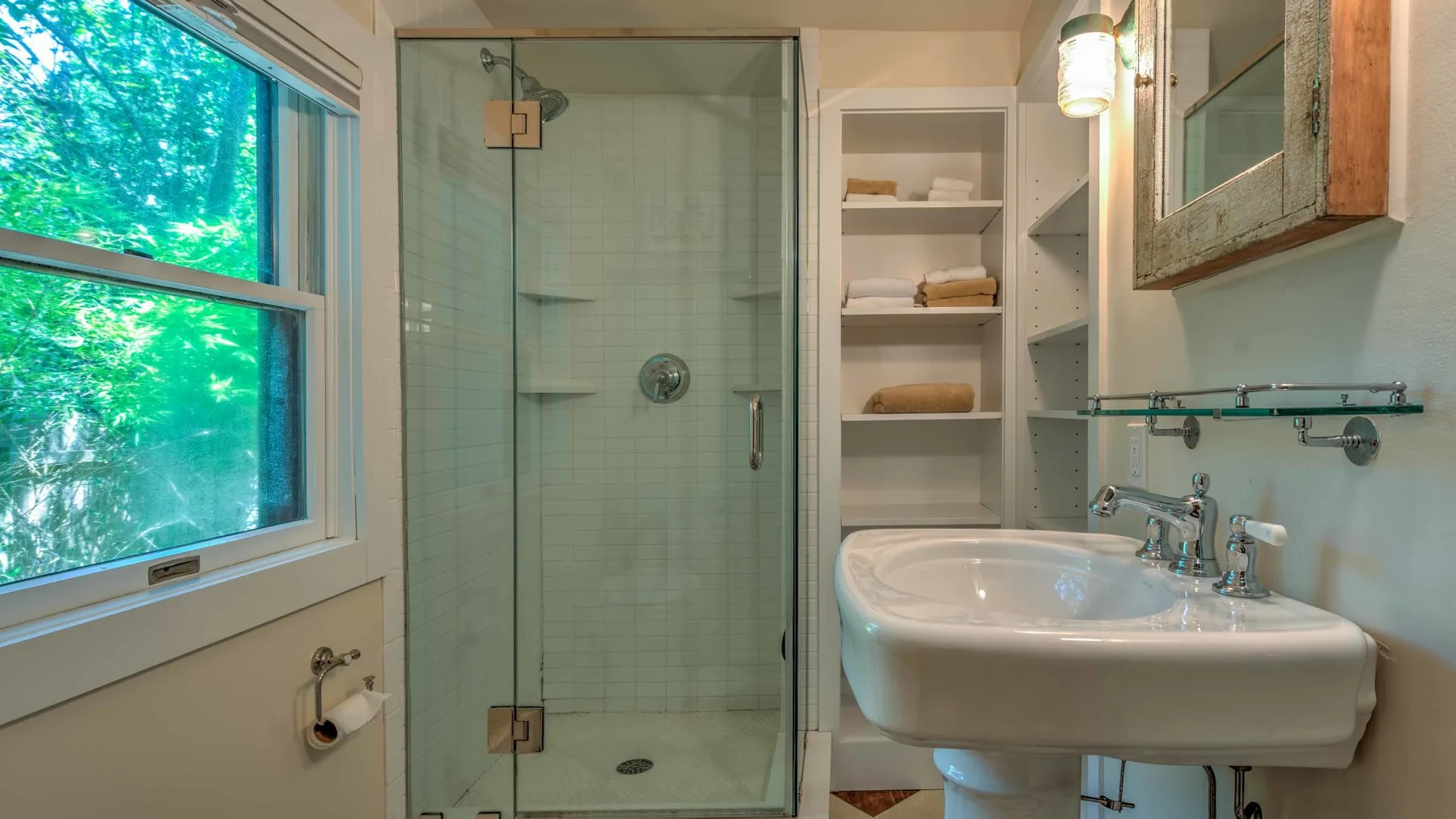 Un baño con ducha de vidrio, lavabo y una ventana con vista a árboles verdes.