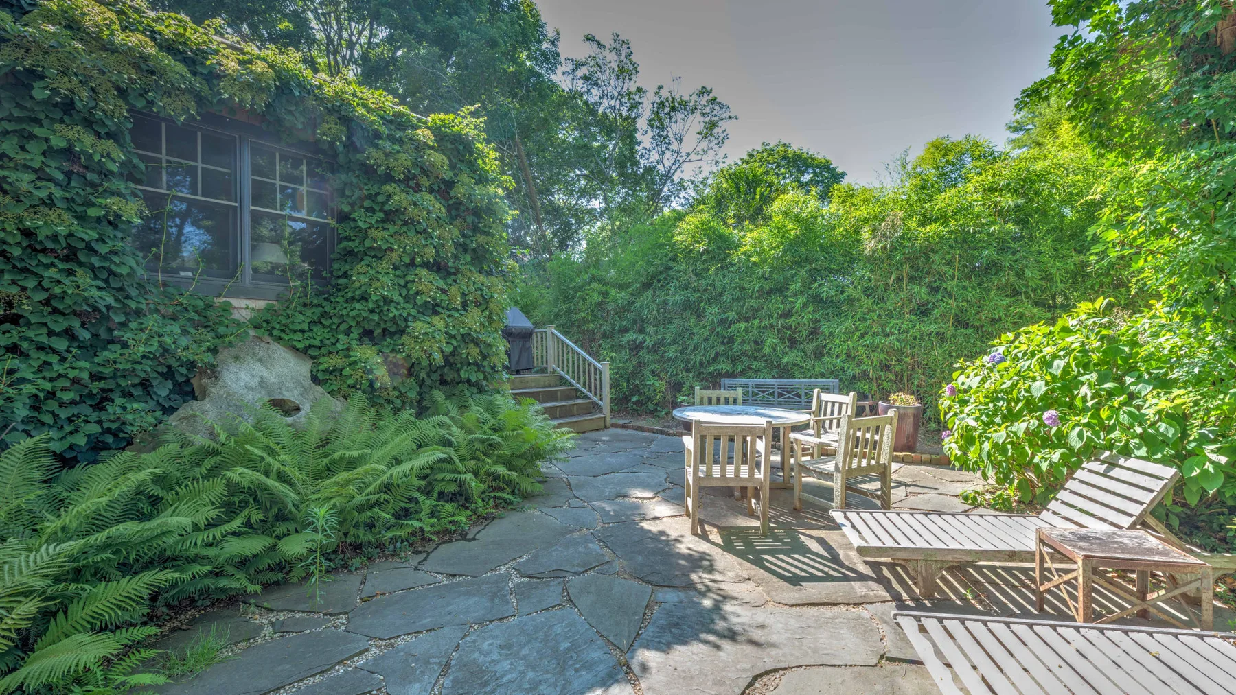 A garden patio with outdoor seating sits among lush greenery and stone paving.