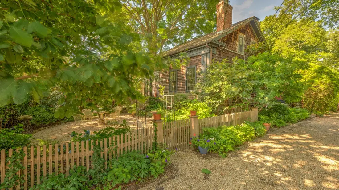 Una casa rústica de madera está entre árboles frondosos, junto a una cerca de jardín.
