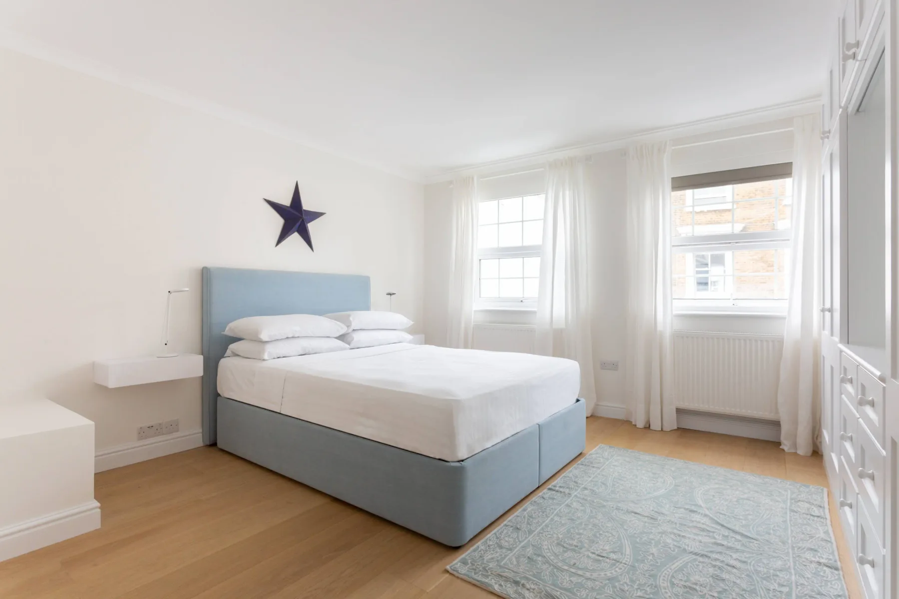 Un dormitorio luminoso con una cama, mesitas de noche y ventanas altas.