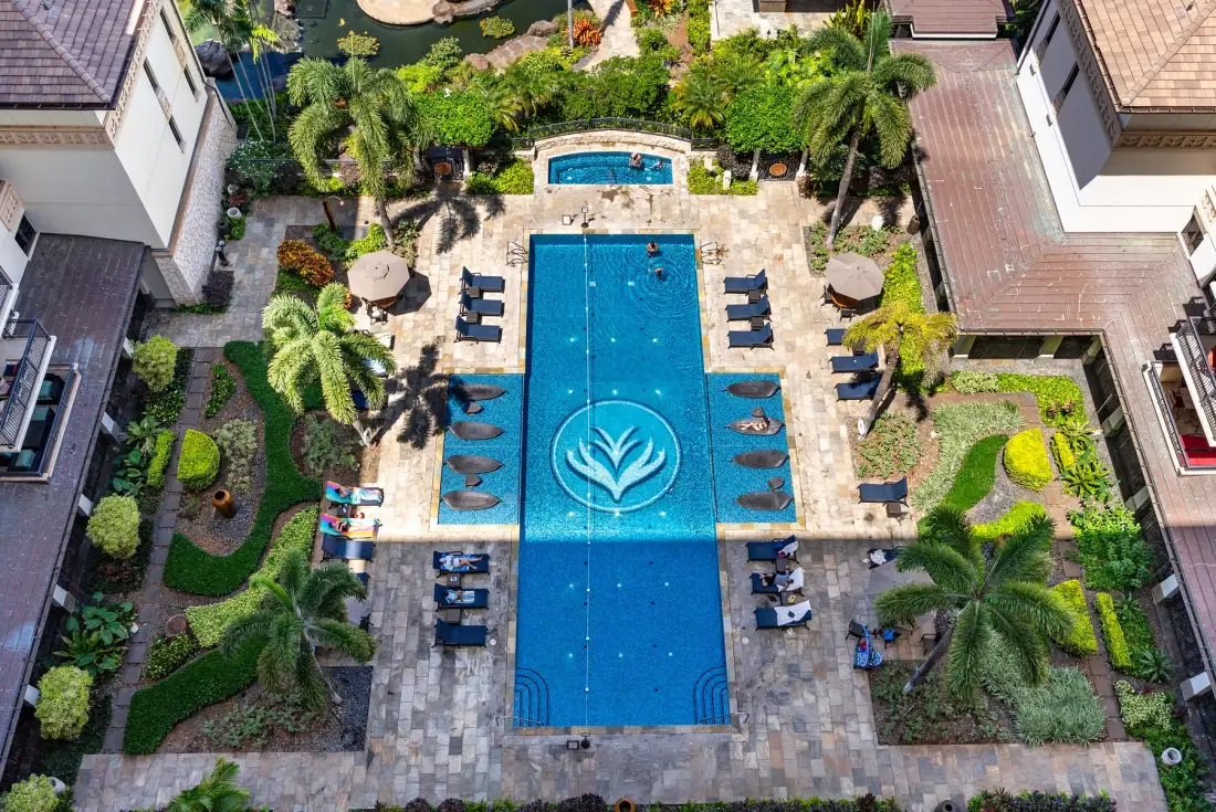Una piscina al aire libre está rodeada de caminos pavimentados, asientos y vegetación tropical.