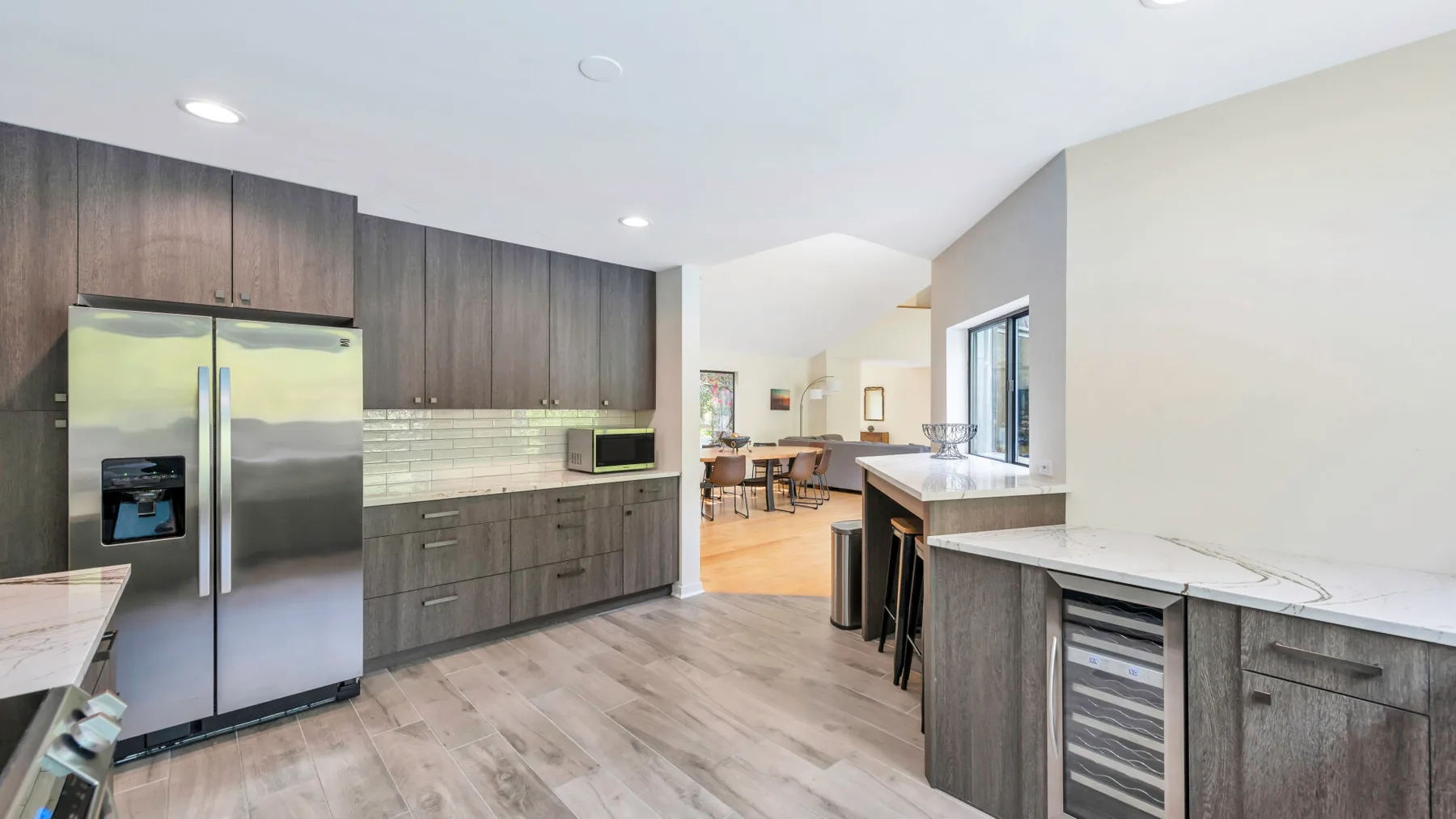 A modern kitchen with dark cabinetry, stainless steel appliances, and a dining area beyond.