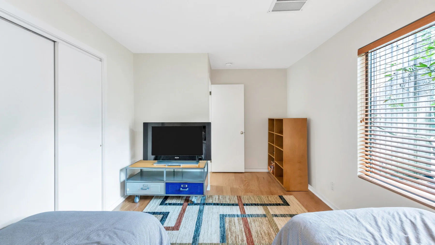 A bright bedroom with a television, wooden shelving, and a large window.
