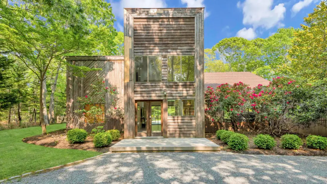 A modern house stands among green trees, shrubs, and a lawn.