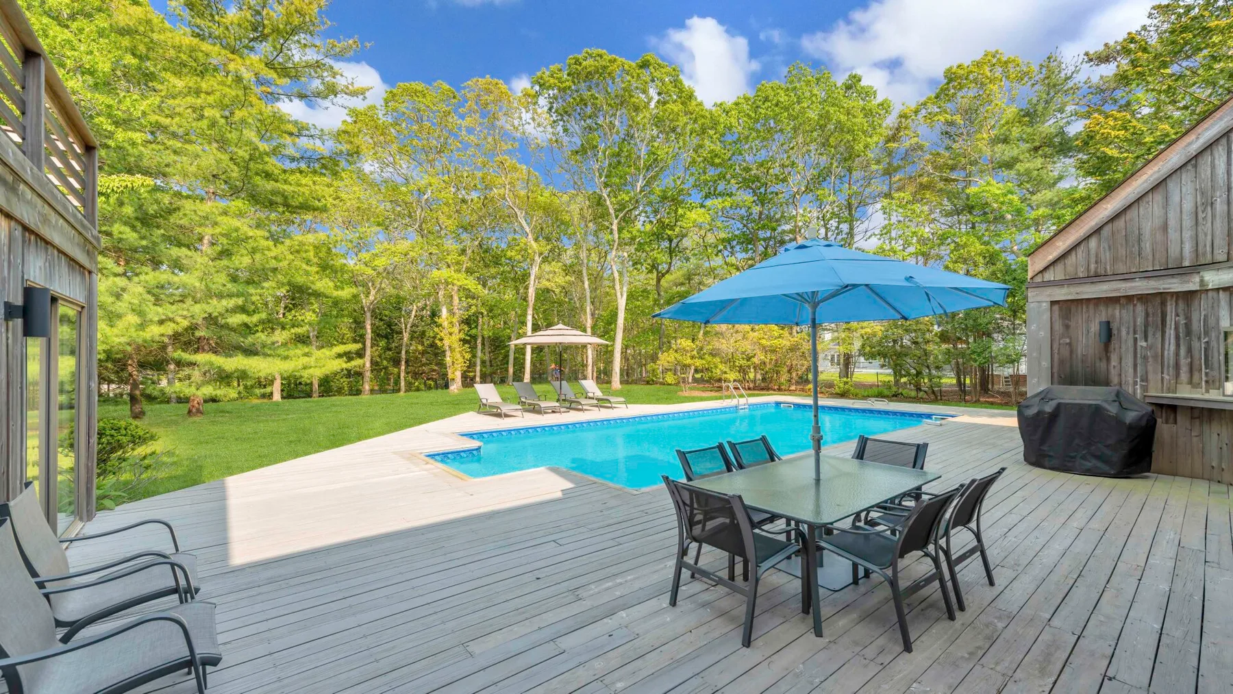 An outdoor pool sits beside a deck with a table and chairs under a blue umbrella.