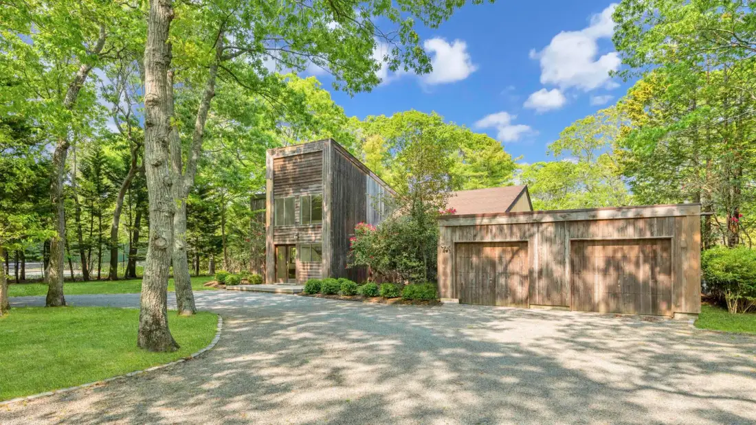 A modern house and garage sit beside a paved driveway among leafy trees.