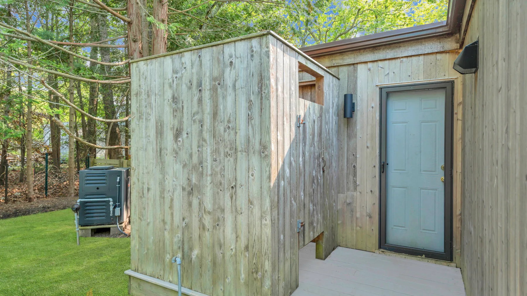 A wood-sided outdoor entry with a gray door beside a grassy, tree-lined yard.