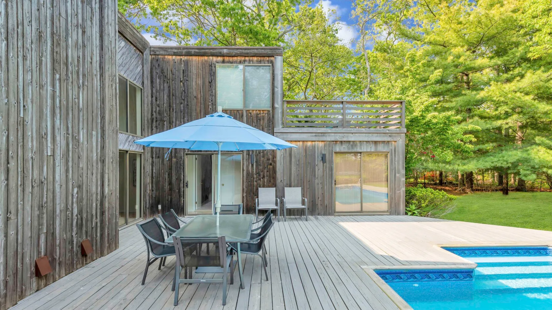 A wooden deck beside a swimming pool has an outdoor dining table and blue umbrella.