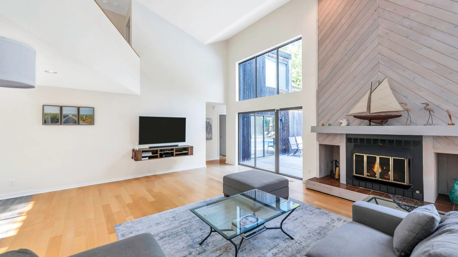 A bright living room has tall windows, a glass coffee table, and a fireplace.