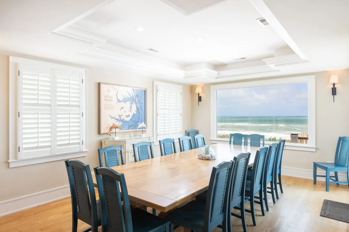 A bright dining room with a long table and a wide window overlooking the ocean.