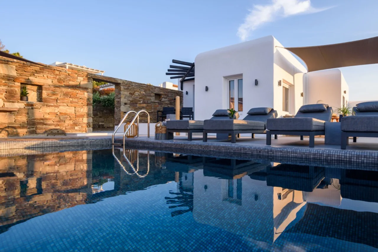 A swimming pool reflects a white villa and outdoor seating in the sunlight.