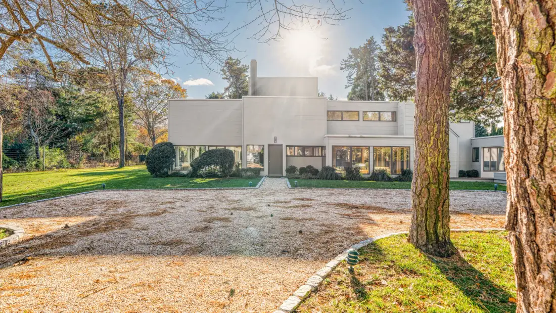 Una casa moderna se alza tras un camino de grava, rodeada de árboles y césped.