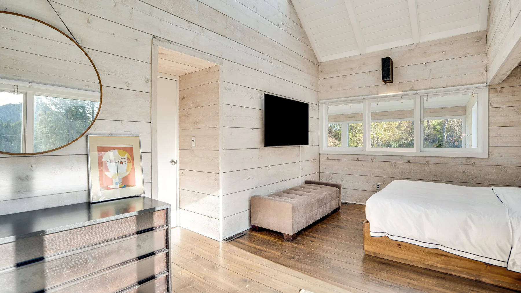 Un dormitorio luminoso con una cama, un asiento tapizado pequeño y un televisor en la pared.