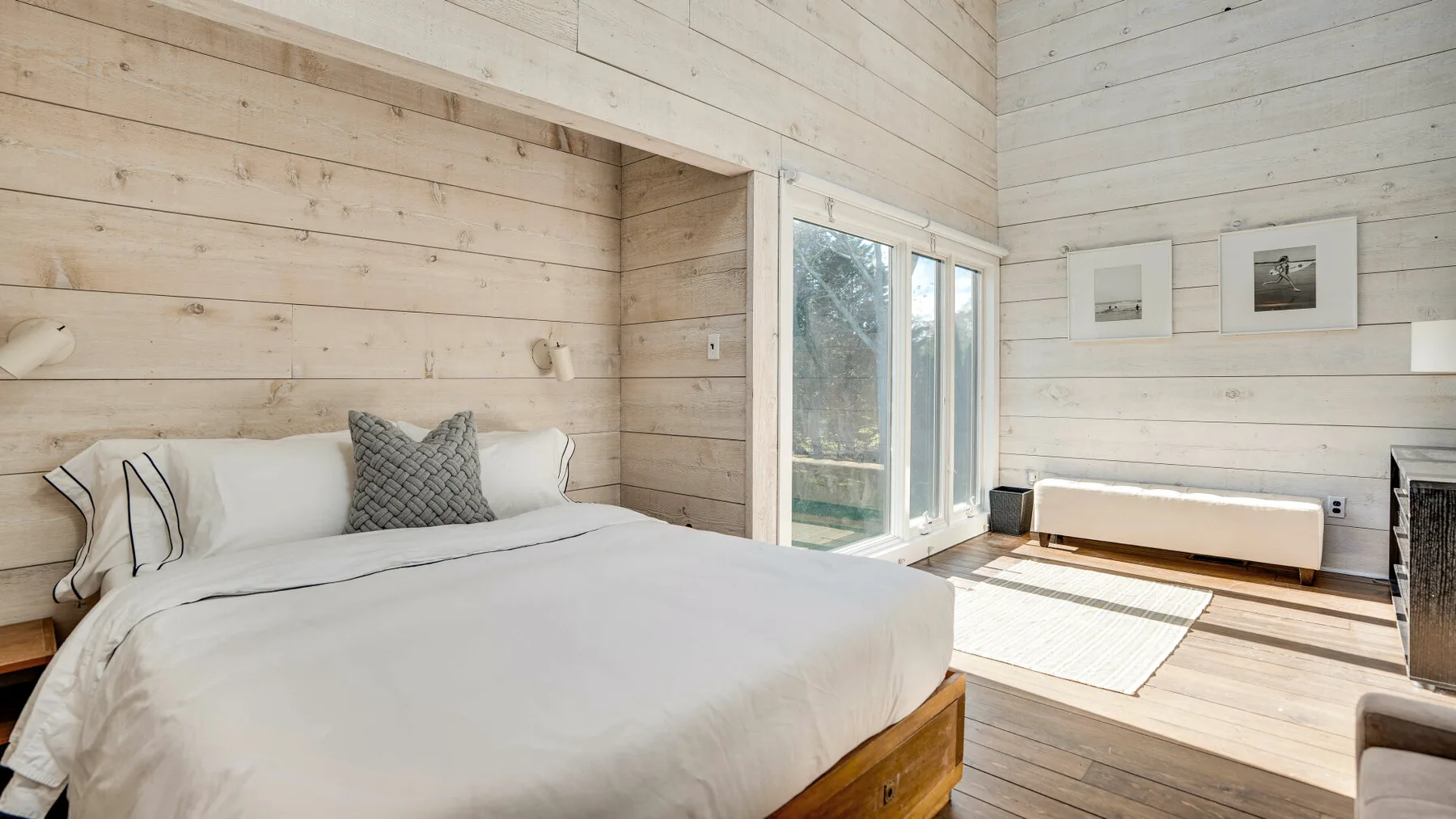 Un dormitorio luminoso con una cama junto a ventanas altas y paredes de madera clara.