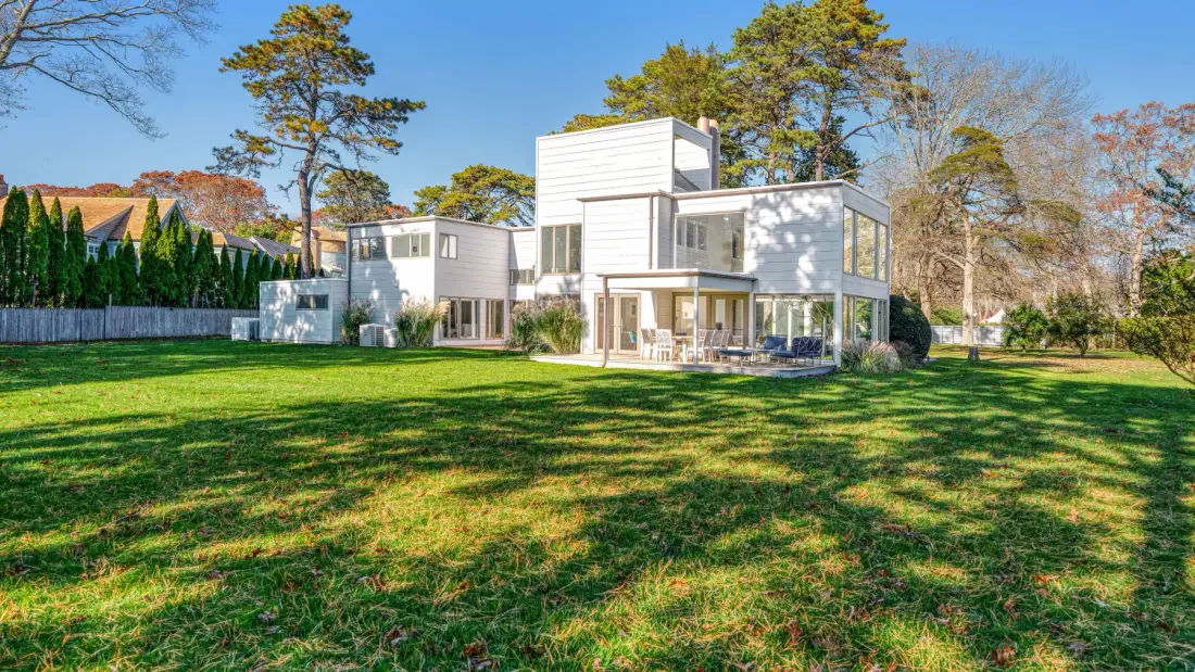 Una casa blanca moderna se alza junto a un amplio césped y árboles maduros.
