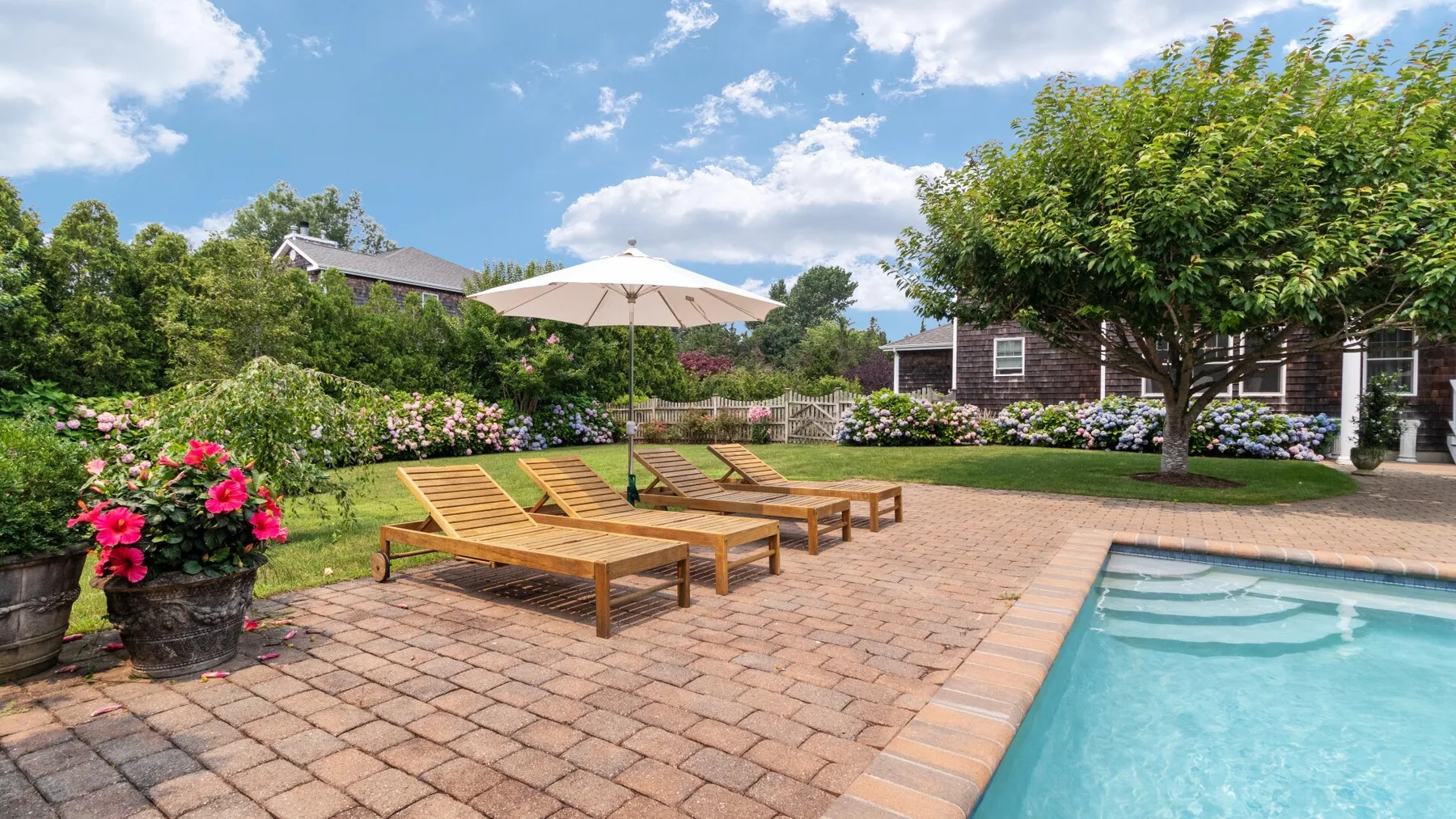 An outdoor pool beside a paved patio with wooden loungers and a shade umbrella.