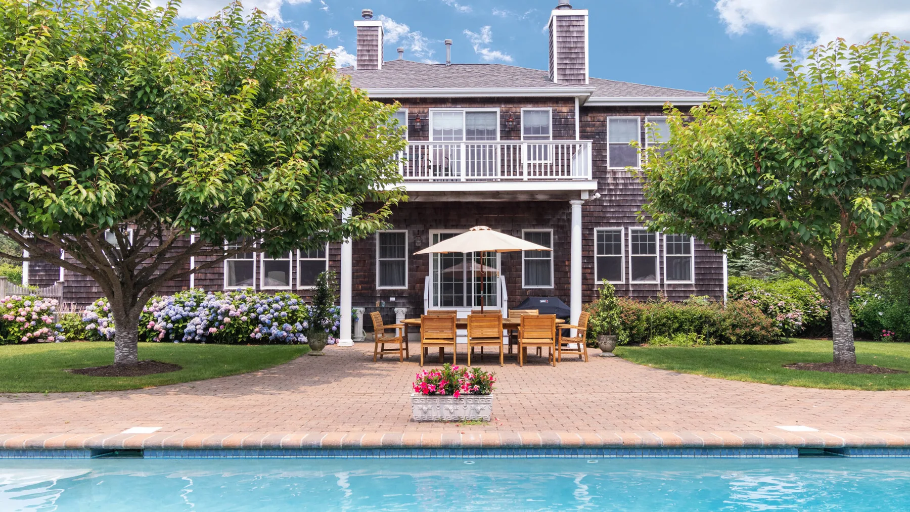 A two-story house overlooks a swimming pool, with patio seating and trees in front.
