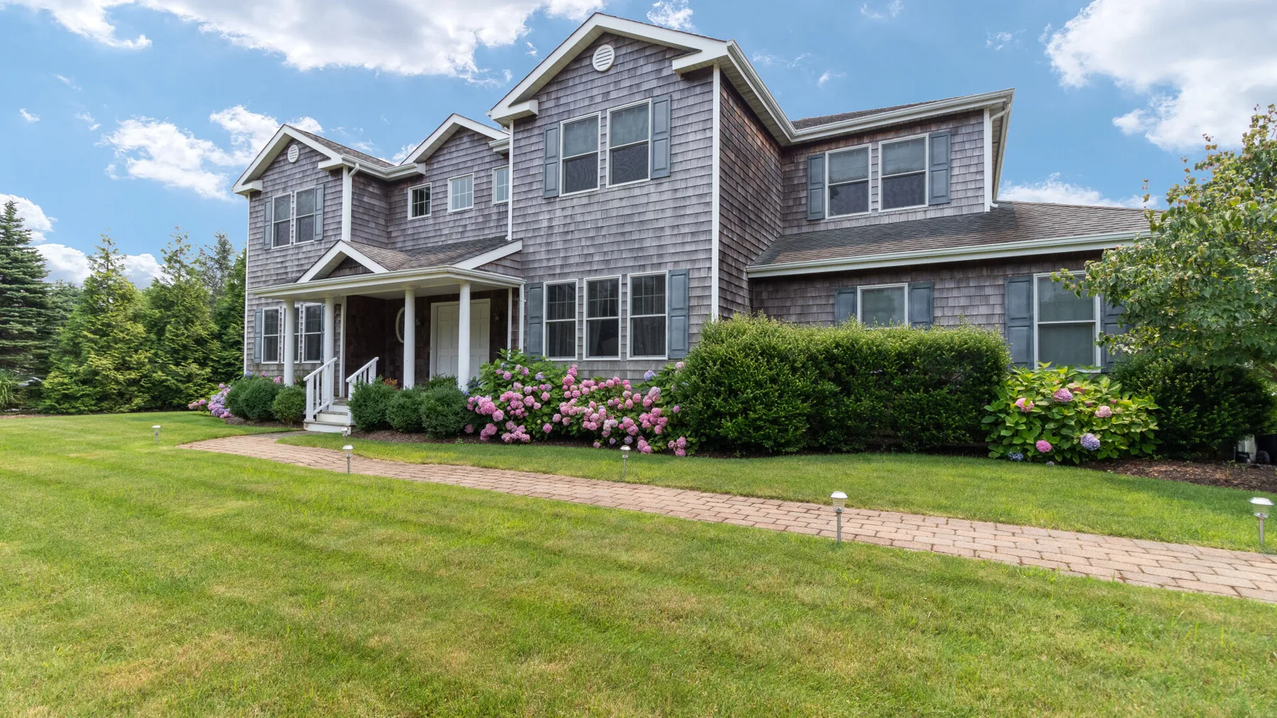 A large two-story house stands behind a green lawn and flowering shrubs.