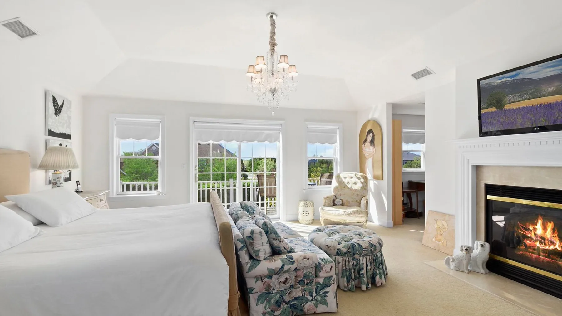 A bright bedroom with a large bed, floral seating, and a fireplace.