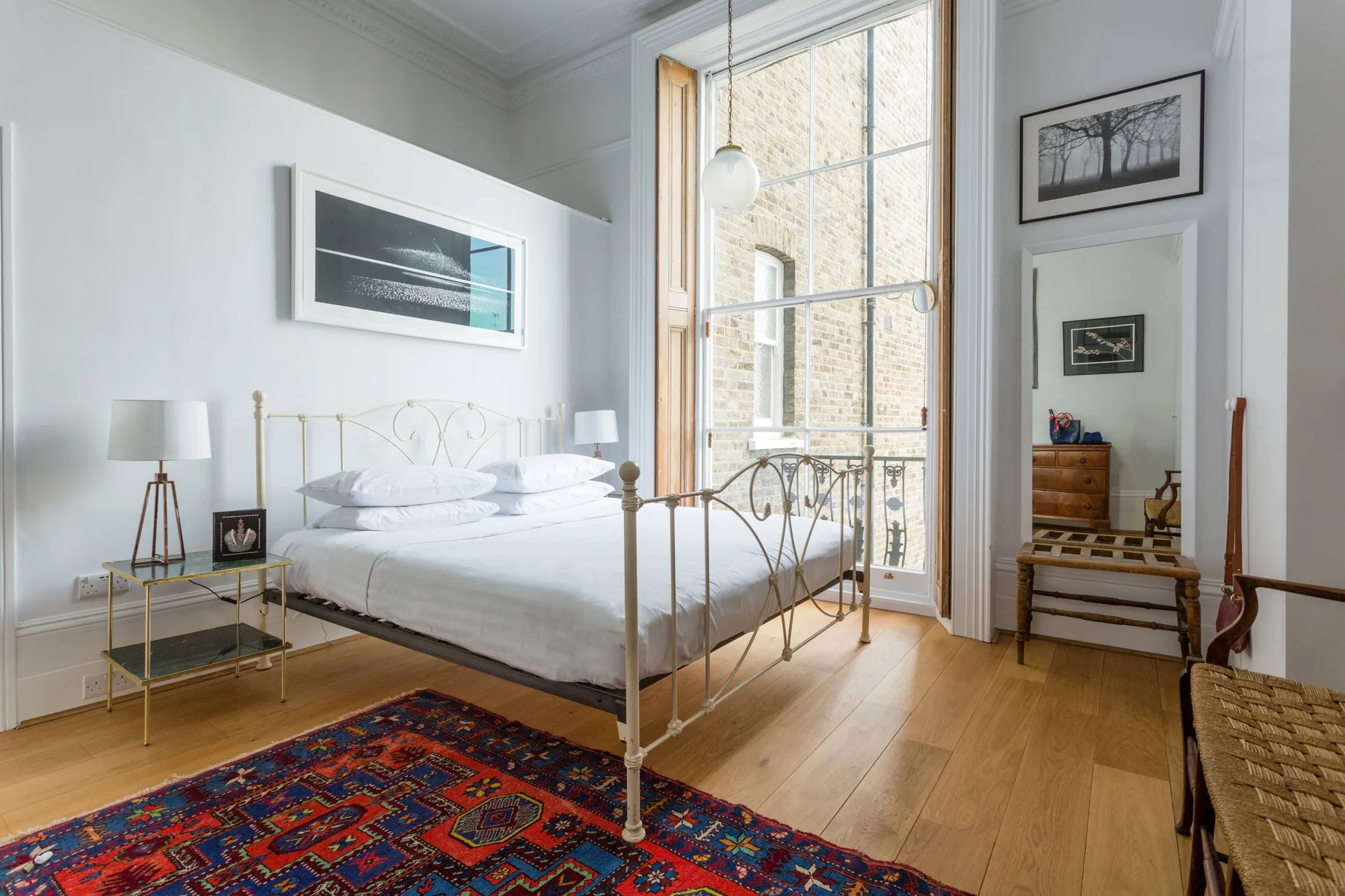 A bright bedroom with a brass bed, tall windows, and a patterned rug.