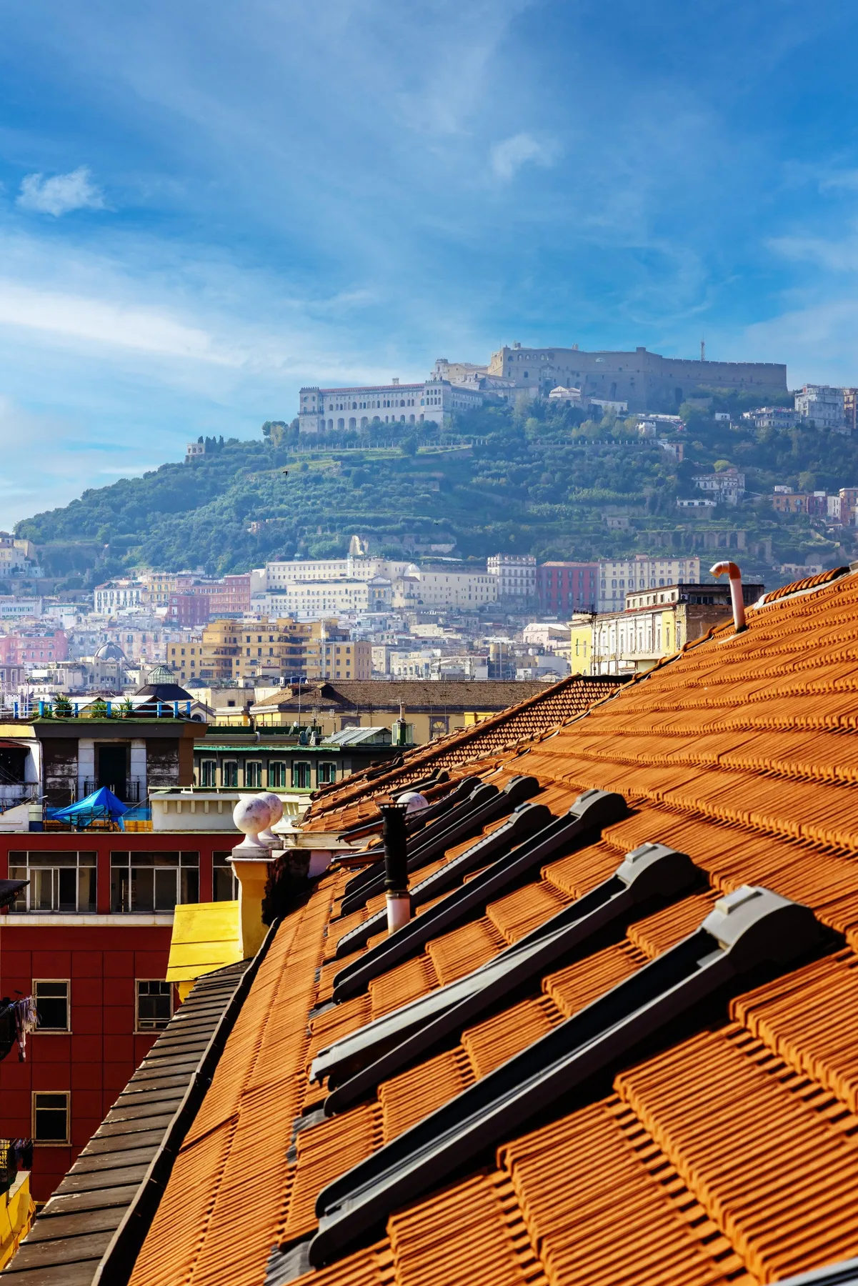 A cityscape with orange tiled rooftops and a hilltop fortress in the distance.