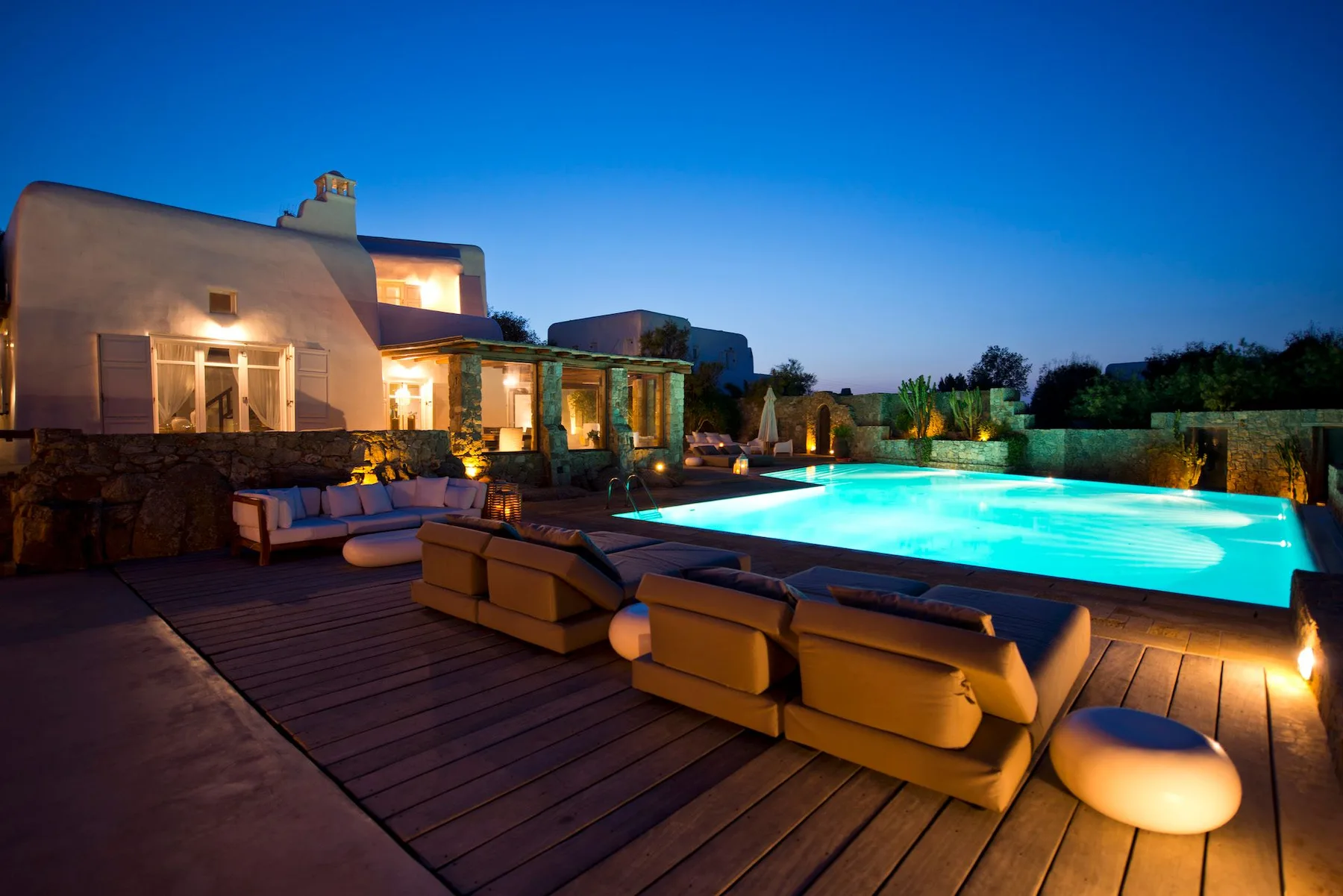 A villa patio with outdoor seating beside an illuminated swimming pool at dusk.