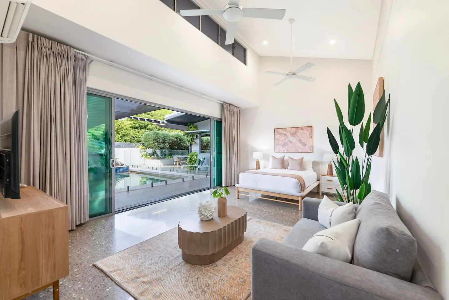 A bright bedroom and seating area open through sliding glass doors to a pool.