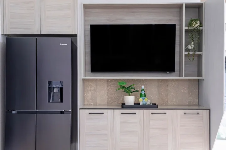 Una cocina moderna con refrigerador, armarios claros y un televisor montado en la pared.