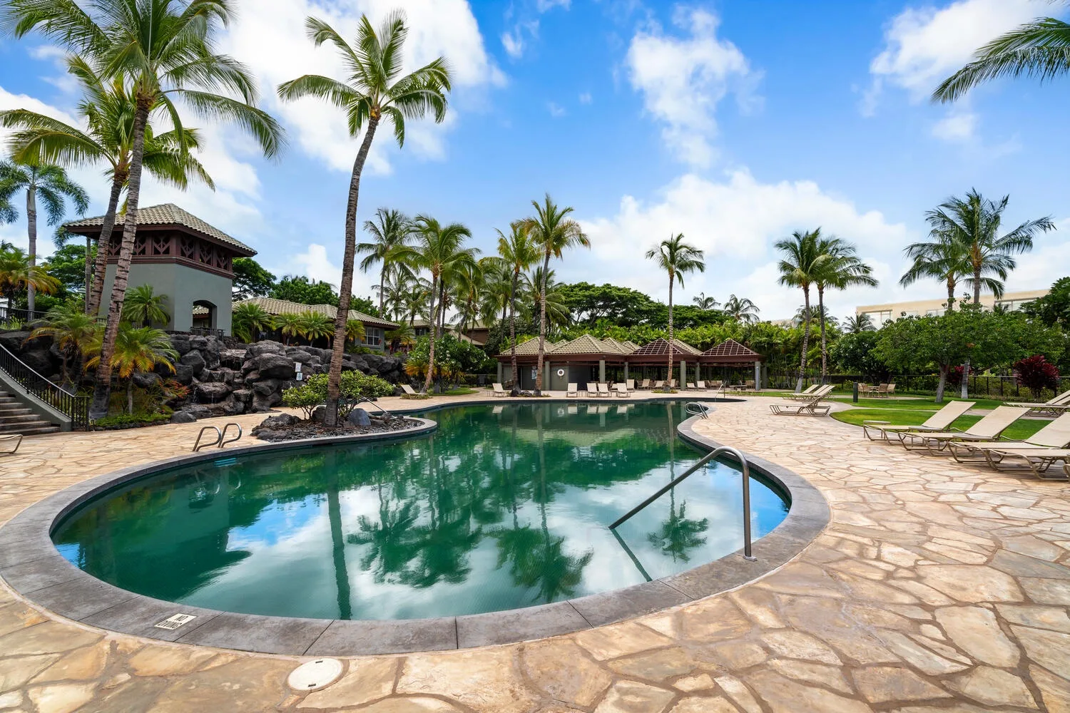 Una piscina tropical al aire libre está rodeada de palmeras, pavimento de piedra y tumbonas.