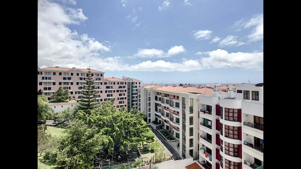 Apartment buildings overlook a tree-lined courtyard beneath a bright, cloudy sky.