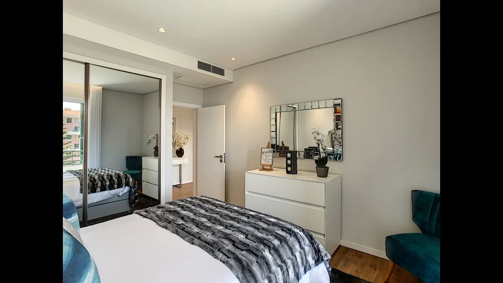 A bedroom with a bed, mirrored dresser, and teal seating.