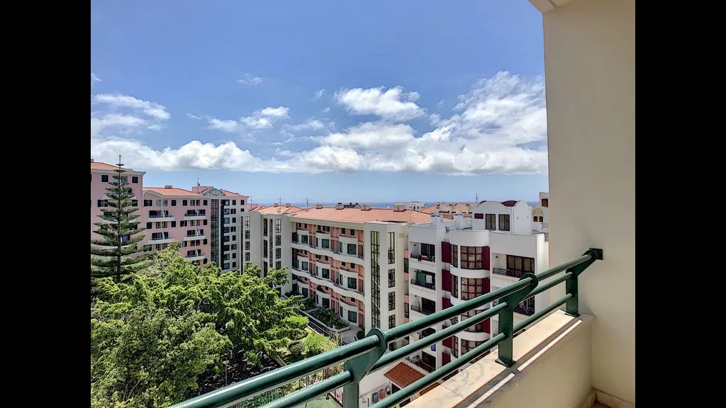 A balcony overlooks nearby buildings, treetops, and the ocean beneath a partly cloudy sky.