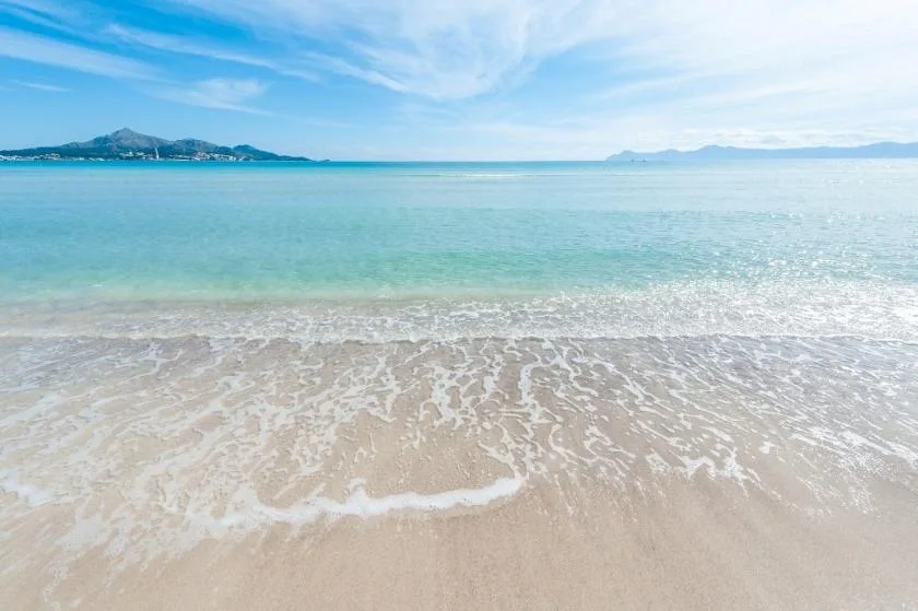 A sandy beach with gentle waves and distant hills across the water.