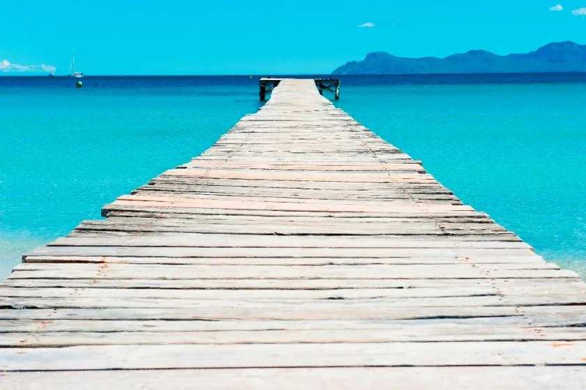 A wooden pier extends over turquoise water toward distant mountains.