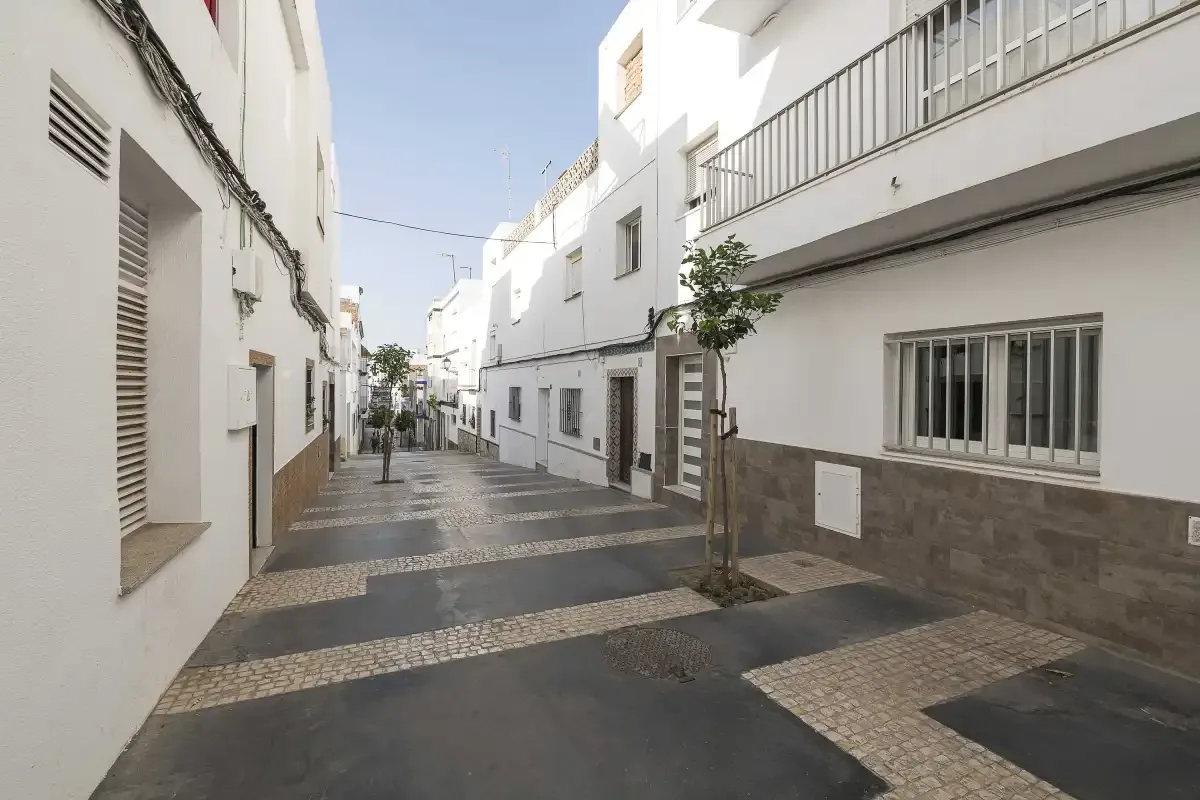 A narrow paved street runs between white buildings, with a small tree beside the walkway.